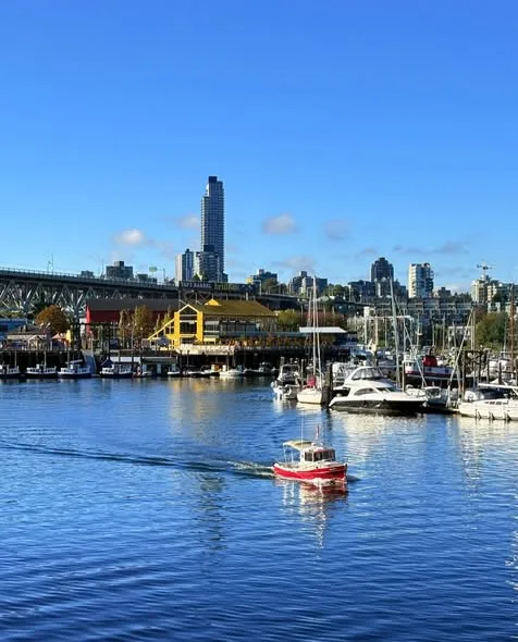 A red motorboat cuts across calm blue water past a busy marina, with downtown Vancouver towers and a bridge behind