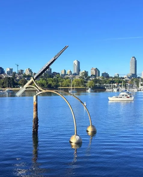 A curved metal sculpture rises from calm blue water, with a sailboat and the city skyline in the distance