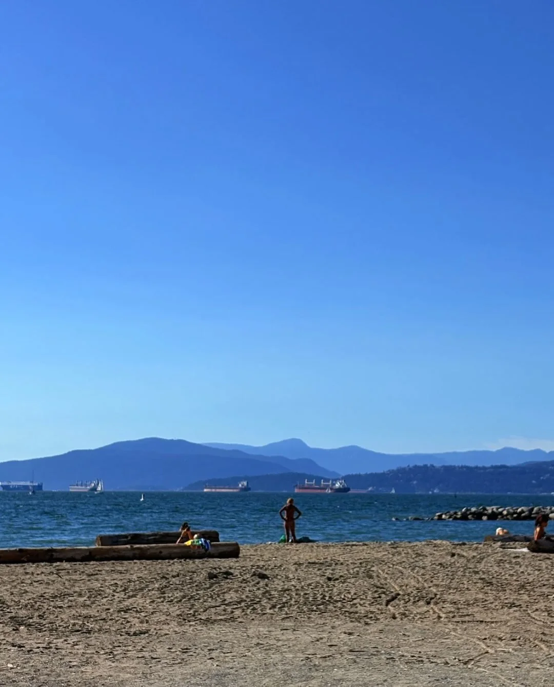 A sandy beach and calm sea stretch toward the North Shore mountains under a clear blue sky